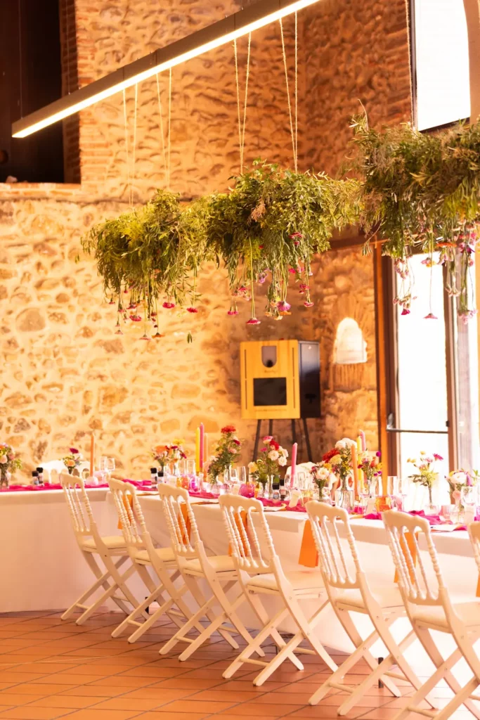 Salle de mariage avec des tables banquets et des suspensions florales colorées dans des tons oranges roses et jaunes.